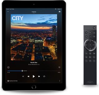 Tablet wyświetlająca oświetlone nocą miasto, obok czarnego pilota Naim Audio NSC 222 z pokrętłem i przyciskami