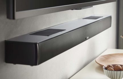 Obdélníkový černý soundbar Canton připevněný na zdi pod televizorem, čelní strana s jemnou kovovou mřížkou