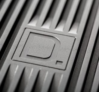 Nærbillede af det reliefprægede "D."-logo på en mat, sort, tekstureret plastikoverfladen af en Definitive Technology BP9060 gulvhøjttaler