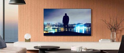 Černý televizor s tenkým rámem, upevněný na světlé dřevěné stěně, zobrazující siluetu před noční panoramatickou linií města, pohled zepředu