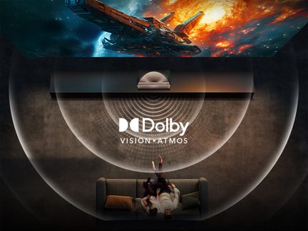 Tmavý salón z pohledu shora, šedá látková pohovka se dvěma osobami, bílý projektor na polici, logo Dolby Vision Atmos