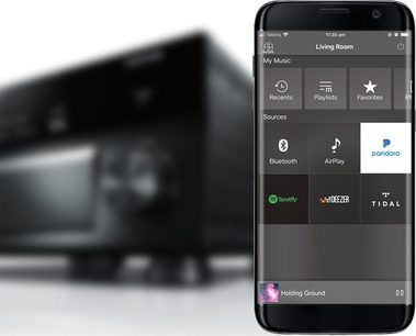 Smartphone, der viser Pandora-appen i en "Living Room"-menu, der lister forskellige lydkilder såsom Spotify, AirPlay og Bluetooth, med en uskarp lastbil i baggrunden