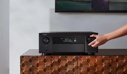 Juodas „Denon“ namų kino stiprintuvas su „Blu-ray“ ekranu ir „Dolby Atmos“, ranka, reguliuojanti garsumo ratuką, ant medinio baldo