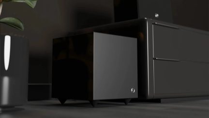 Kompakter schwarzer Subwoofer mit dunkelgrauer Front und schräg gestellten Füßen, in Dreiviertelansicht