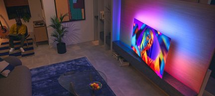 Large black television with a slim matte-finish frame mounted on a light wood wall panel, multicolored backlight halo, seen from an oblique overhead angle