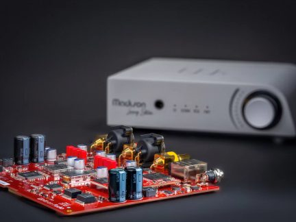 Red audio circuit board with connectors and capacitors in front of a white Mola Mola amplifier