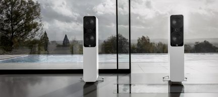 Pair of white floorstanding speakers with a lacquered finish and black front panel, two round drivers and a centrally aligned tweeter in a vertical arrangement, front view, placed on metal stands in front of a bay window
