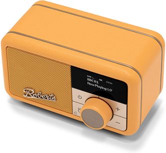 Roberts Revival Petite 2 gul bordradio, kompakt rektangulært kabinet i kornet kunstlæder med metalkanter, set fra tre kvart forfra i let fugleperspektiv. Fronten består af et perforeret metalgitter til venstre med det kromede logo, et sort display til højre og en stor cirkulær drejeknap i beige omgivet af plus- og minus-knapper.