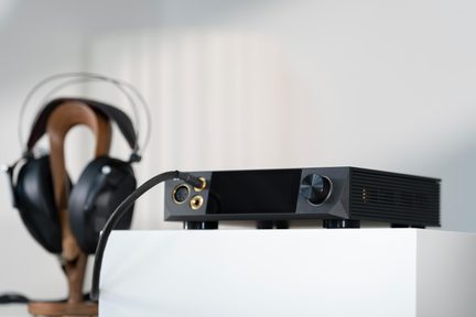 fekete hi-fi fejhallgató-erősítő nagy gombbal és aranyozott audio csatlakozókkal, fa állványra helyezett fülkagylót körülölelő fejhallgató hátulról