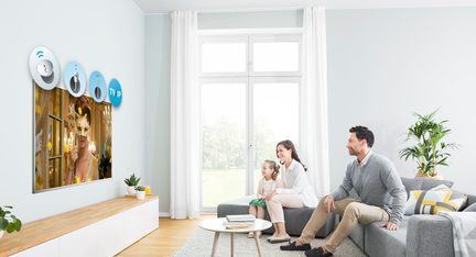 Rodzina trzech osób siedząca na szarej kanapie w salonie z dużymi oknami i telewizorem naściennym wyświetlającym maski