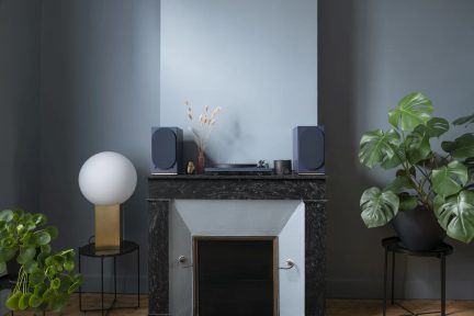 Woonkamer vooraanzicht, schoorsteen in zwart marmer, platenspeler en twee blauwe luidsprekers, witte bolvormige lamp en groene planten