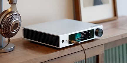 Audioverstärker aus mattiertem Aluminium in hellem Farbton, Dreiviertelansicht von vorne links, Front mit zentralem Farbdisplay, großem schwarzem Drehregler und 6,3-mm-Kopfhöreranschluss, linke Seite mit schwarzem Lüftungsgitter