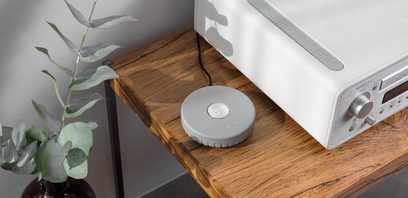 Netværksaudioafspiller Audio Pro Link 1 i lys grå på træhylde, set ovenfra i vinkel, grøn plante til venstre