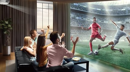 Vedere din spate, din unghi de trei sferturi, a unui living contemporan, cu un videoproiector alb montat pe tavan care proiectează un meci de fotbal pe un ecran mare, în fața unei canapele de colț negre din piele ocupate de mai mulți spectatori entuziaști