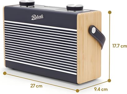 Rádio Roberts Rambler Max z pohledu tří čtvrtin zepředu vpravo, obdélníkový pouzdro z tmavě modrého plastu s přední stranou s horizontálními bílými pruhy, boky z lakovaného světlého dřeva, dva velké kulaté kovové knoflíky na horní straně a postranní řemínek z tmavě modré kůže s dekorativním šitím.