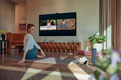 Kvinde siddende på et trægulv i en stue, ser på et tv-skærmbillede, der viser yoga- eller fitnessbilleder