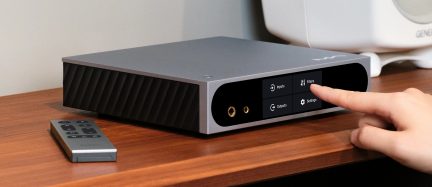 Sudraba alumīnija audio korpuss, trīs ceturtdaļu skats no priekšpuses, melna spīdīga priekšpuse ar skārienekrānu, uz kura ir četras ikonas un divas zeltītas austiņu ligzdas, kreisajā sānā slīpas ventilācijas atveres.