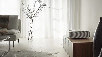 Moderní minimalistický interiér s šedou pohovkou, dřevěným konferenčním stolkem, vázou s větví a projektorem Samsung The Premiere SP-LSP9TFA
