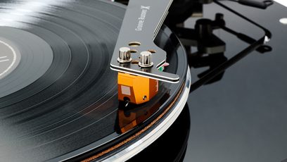 Vista cercana oblicua de un tocadiscos hi-fi Vertere Acoustics Pack DGX Sabre blanco, con base de acrílico negro brillante y plato de aluminio que sostiene un disco de vinilo negro, brazo de lectura metálico Groove Runner X terminado en una cápsula naranja fijada con dos tornillos plateados.