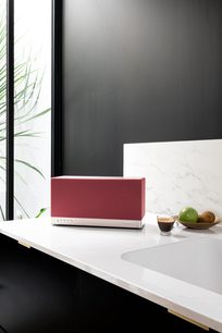 Triangle AIO 3 connected speaker in ochre red, front view on a white countertop with a mug and fruits
