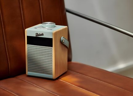 Radio portatile rettangolare vista da tre quarti frontale, custodia in impiallacciatura di legno chiaro con rifiniture in pelle blu chiaro, griglia frontale perforata in metallo argentato, grande pulsante rotondo metallico sulla parte superiore, piccola cinghia laterale coordinata