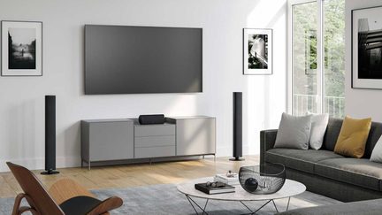 Moderne stue med kæmpestor væghængt tv-skærm, gråt tv-møbel, sorte gulvhøjttalere og grå sofa med farverige puder