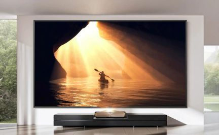 Pie sienas piestiprināts lielformāta televizors, kurā redzams kajakotājs saules staru apgaismotā alā
