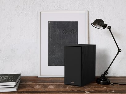 Sort trådløs Klipsch R-40PM højttaler, set forfra, med teknisk diagram på hvid ramme og sort skrivebordslampe