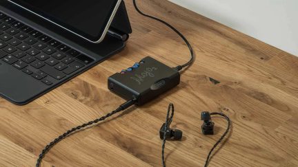 Let, let oppefra af et lille, rektangulært sort audiokabinet i mat aluminium, forbundet med kabel til en sort tablet på tastatur, med fire farvede, kugleformede knapper på toppen og placeret på et lyst træskrivebord ved siden af et par sorte in-ear-hovedtelefoner med snoet kabel