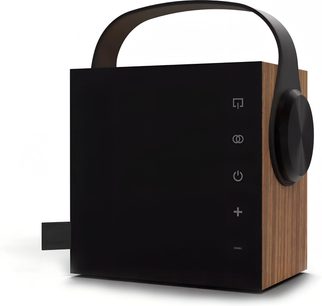 Bärbar bluetooth-högtalare Morel Biggie ek, sedd snett framifrån höger, kubformad låda med sidor i ekfaner och matt svart front, utrustad med ett svart bågformat handtag upptill, ett stort runt svart vred på högra sidan och vertikalt uppradade touchkontrollikoner.