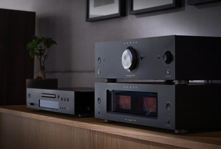 Černá hi-fi sestava z kovu, zesilovač s velkým ovladačem a VU metry, přehrávač se šuplíkem, pohled ze tří čtvrtin na dřevěném nábytku