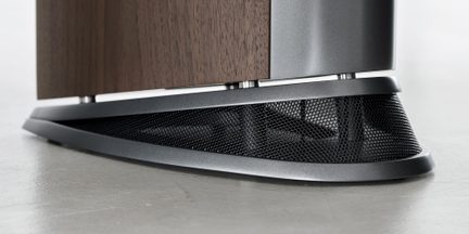 Nærbillede fra siden i frøperspektiv af en skråtstillet oval base i gråt metal med sort perforeret gitter, der understøtter et lodret panel i brunt træfinér på et gråt gulv