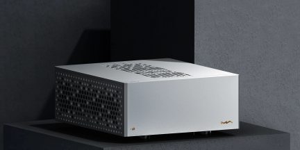 Silbernes Elektronikgehäuse aus gebürstetem Aluminium, Dreiviertelansicht von vorne links in leichter Aufsicht, Oberseite und linke Seite mit runden Lüftungsöffnungen versehen, goldene runde Kontrollleuchte unten an der Front und dezentes goldenes Logo