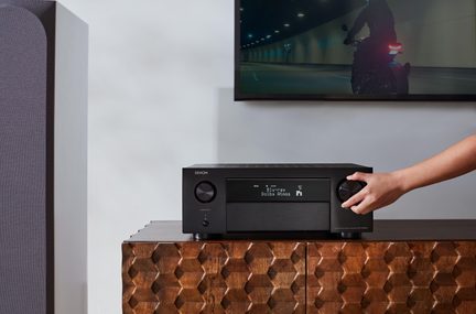 Schwarzer Denon AV-Verstärker auf Holzmöbel, Lautstärkeregler von Hand eingestellt, Fernseher im Hintergrund eingeschaltet