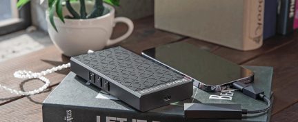 Custodia rettangolare nera con texture, con 4 porte visibili, cavo collegato, smartphone nero, libro, tazza bianca, pianta verde.