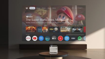White portable projector displaying the Google TV interface with the movie The Super Mario Bros. Movie in a modern living room