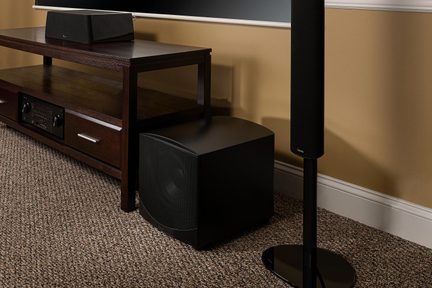 Cassa nera cubica su mobile in legno scuro, vista laterale, con soundbar nera rettangolare davanti