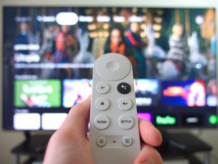 Vit fjärrkontroll hålls i handen, synliga knappar, suddig TV-skärm i bakgrunden med appgränssnitt