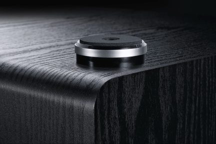 Disc circular negru și argintiu din metal și material texturat, așezat pe un colț al unei suprafețe din lemn negru cu vene vizibile.