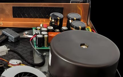 Detailní záběr elektronických součástek, černých válcových kondenzátorů, červených a černých kabelů, černého kruhového pouzdra