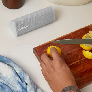 Gray Bluetooth speaker, hand holding a knife cutting a lemon on a wooden cutting board, close-up view