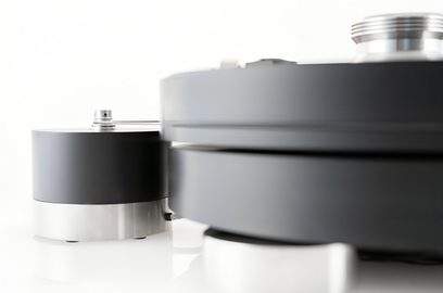 Vista lateral cercana de un tocadiscos hi-fi Transrotor Max Nero RB330 con acabado negro mate, con un gran plato cilíndrico desenfocado en primer plano apoyado sobre un pie de aluminio cepillado y un bloque motor cilíndrico negro con base metálica al fondo