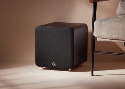 Matte black cubic metal subwoofer with fabric grille, three-quarter front right view, rounded corners, silver circular logo at the bottom, standing on four conical metal feet