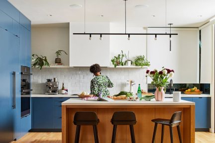 Moderne blåt køkken med træ-køkkenø, grønne planter, sort lineær loftlampe og en person set bagfra, der laver mad
