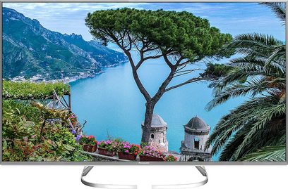 Panasonic TX-50EX730E-fladskærms-tv, der viser et kystlandskab med fyrretræer, tårne og hav, set forfra på sølvfarvet fod