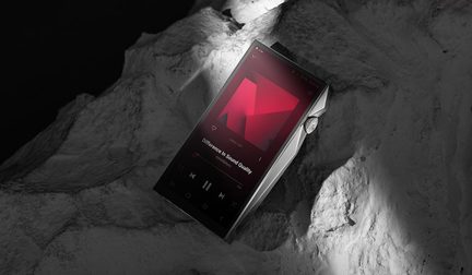 Lettore audio portatile rettangolare in metallo argentato, schermo touch nero con interfaccia rossa, rotella zigrinata sul lato destro, vista a tre quarti