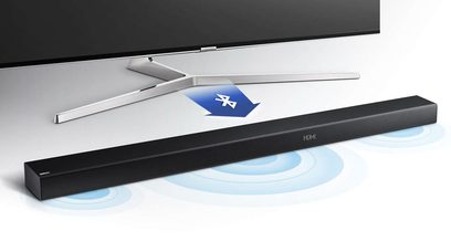 Černý soundbar Samsung HW-M450, pohled shora se stříbrným dálkovým ovladačem položeným na něm, na světle šedém pozadí s modrými odlesky