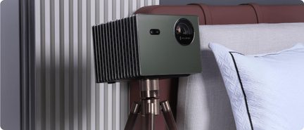 Olive green rectangular metal projector, smooth front with circular lens and sensor, black finned sides, mounted on a metal tripod, three-quarter front view