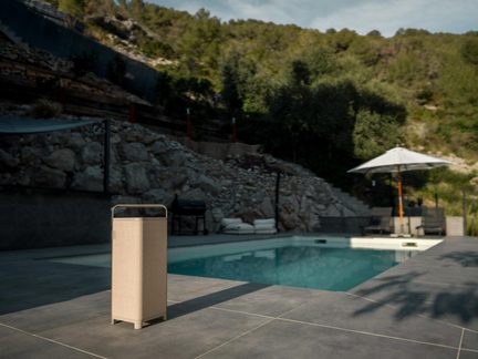 Bärbar beige rektangulär Bluetooth‑högtalare med handtag, placerad på en kaklad terrass nära en pool