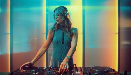 Person i halvfigur, sort og sølvfarvet hjelm, ved sorte metal-DJ-pladespillere med lysende knapper, baggrund af diffuse cyan- og ravfarvede paneler